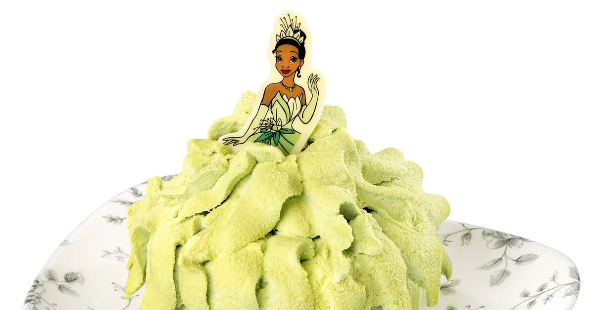 Tiana Kuchen Auf einem Teller wird ein tiana-Kuchen präsentiert. Der Kuchen hat dabei die Form von Tianas hellgrünem Kleid und verjüngt sich nach oben. Dort steckt eine Oblate die Tianas Oberkörper und Gesicht zeigt