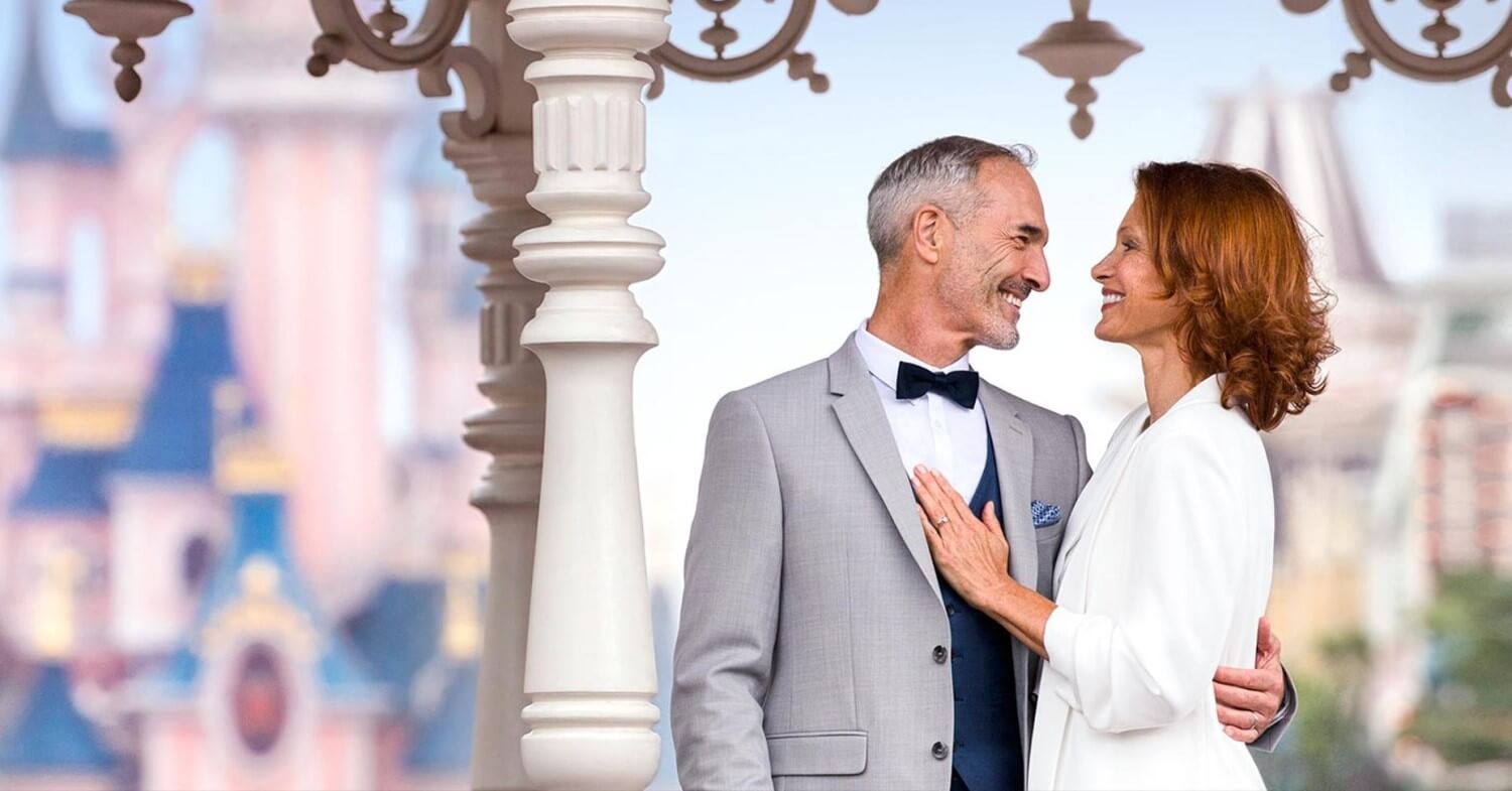 Eheversprechen Disneyland Paris Ein Paar in den besten Jahren schaut sich verliebt in die Augen. Beide tragen festliche Kleidung, im Hintergrund ist das Schloss von Disneyland Paris zu sehen