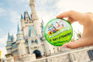 Eine Hand hält einen Button mit der Aufschrift "I'm Celebrating 15 Years of Memories" vor dem Cinderella Castle in die Höhe