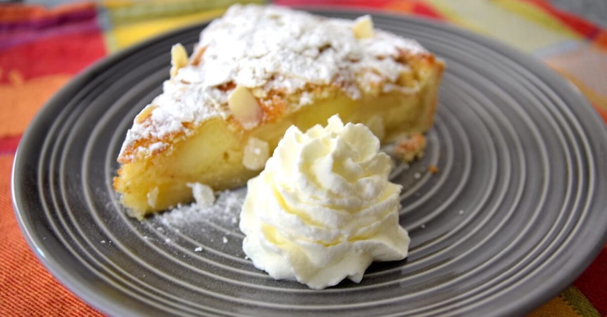 Auf einem grauen Teller ist ein Stück mit Puderzucker bestäubter Apfelkuchen mit Sahne zu sehen