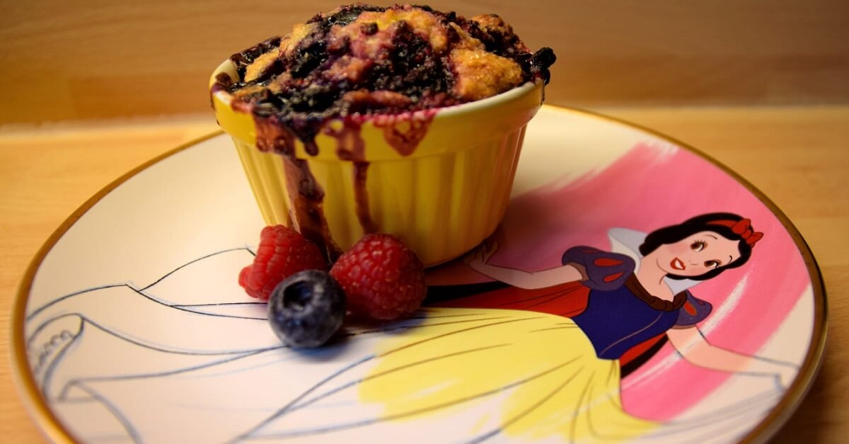 Auf einem Teller mit Schneewittchen-Motiv steht ein Schälchen mit leckerem Berry Cobbler mit einigen frischen Him- und Blaubeeren