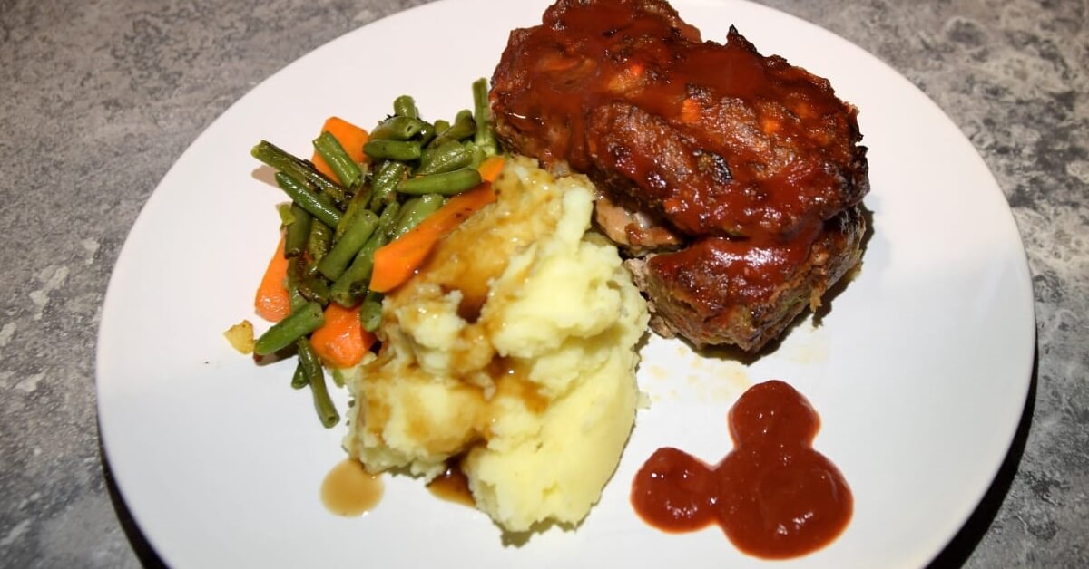 Auf einem großen weißen Teller liegt ein kleiner Hackbraten mit Kartoffelpüree und Gemüsebeilage