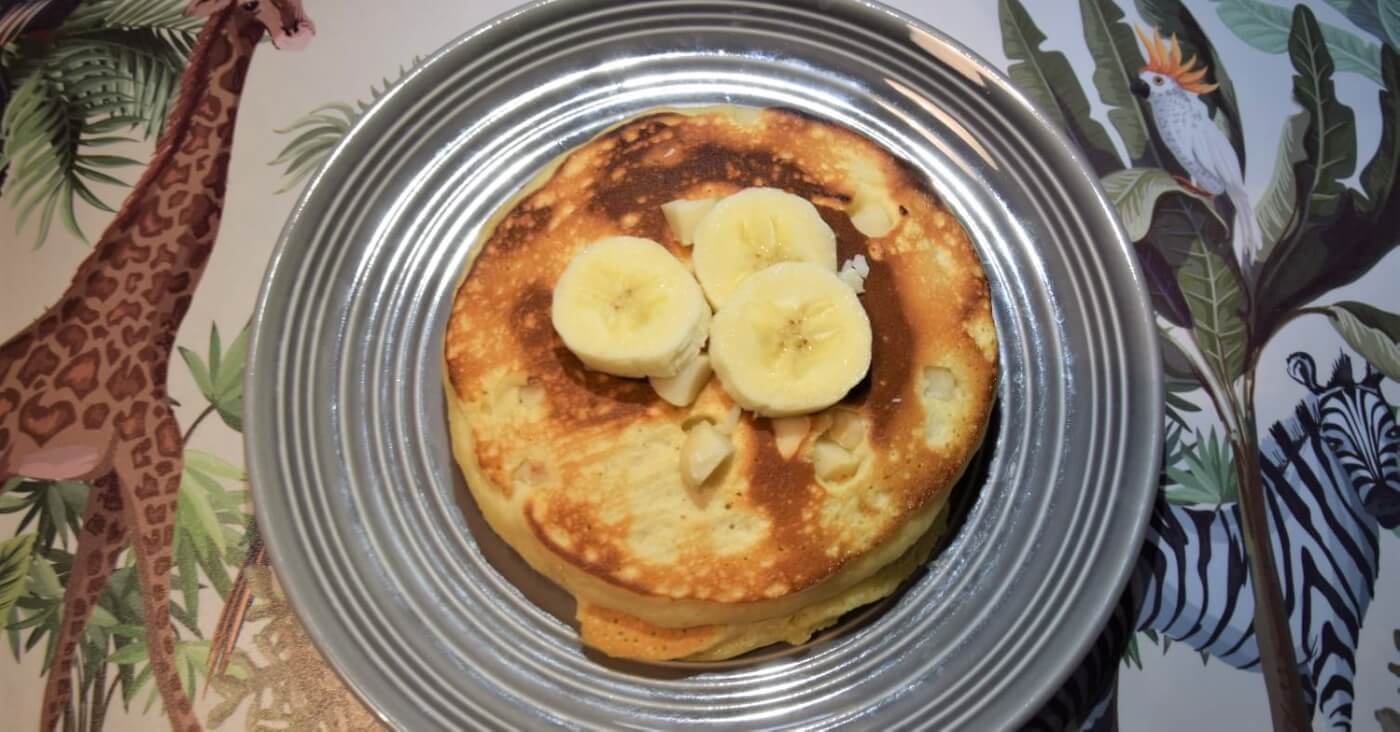 Auf einem grauen Teller liegt ein Stapel von Pancakes mit Macadamianüssen und Bananenscheiben obenauf