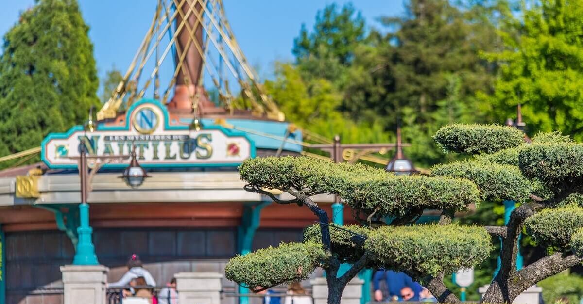 Vor einem Baum mit eckig geschnittenenem Grün vor dem Eingang zur Attraktion Les Mysteres du Nautilus im Discoveryland