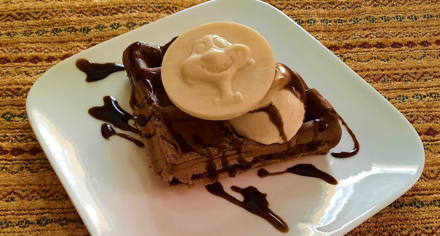 Auf einem kleinen quadratischen weißen Teller liegt eine Schokoladenwaffel mit gefrorener Espresso-Mousse und einem weißem Schokoladentaler mit einer Abbildung von Timon aus Disney's König der Löwen