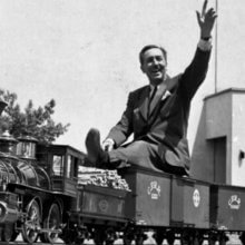 Walt Disney sitzt winkend auf dem Modellzug, der auf seinem Grundstück fährt.