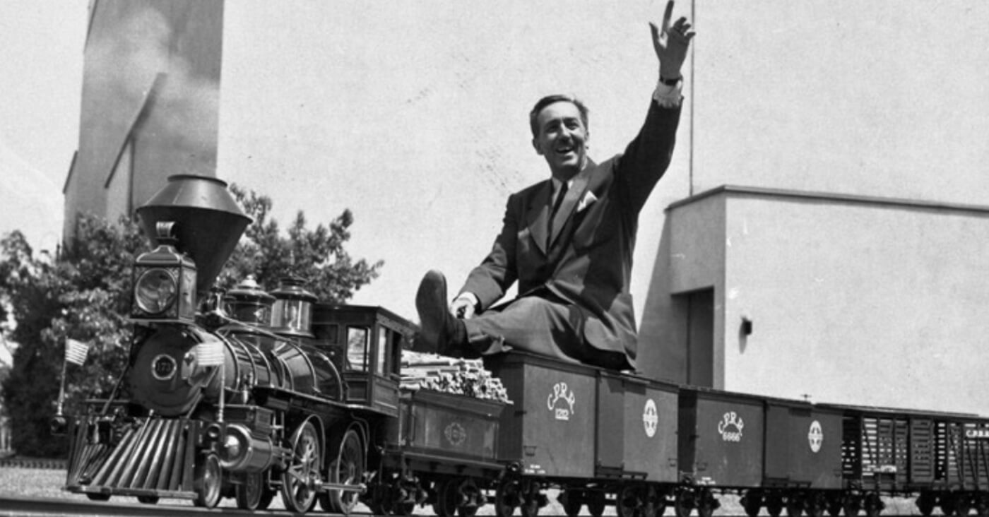 Walt Disney sitzt winkend auf dem Modellzug, der auf seinem Grundstück fährt.