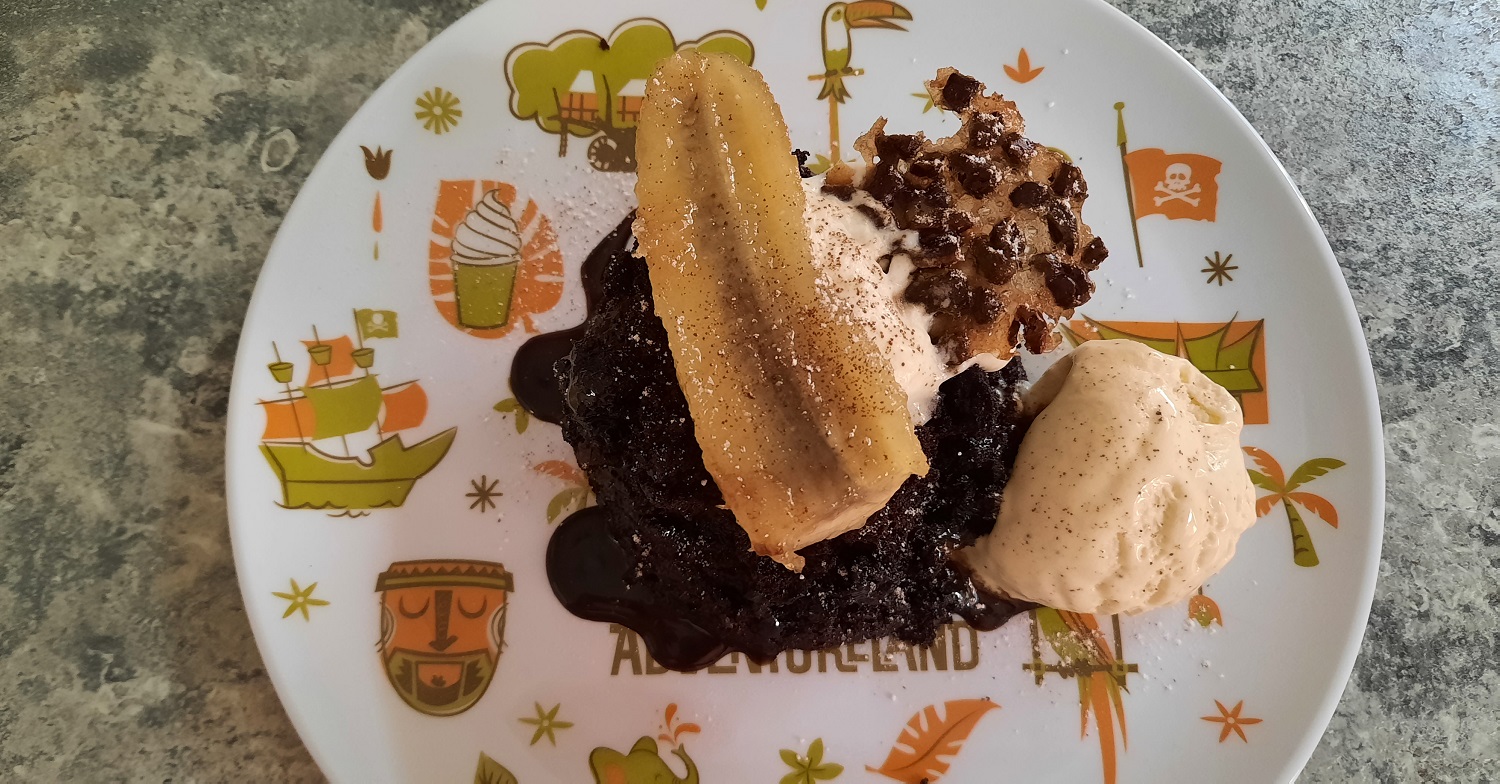 Ein Schokoküchlein mit Schokoladensauce, karamellisierter Banane, Vanilleeis, Sahne und Cacao Nib Tuile auf einem Teller mit Adventurelandmotiv