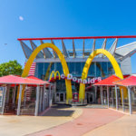 Blick auf ein McDonald's Restaurant mit Glasfassade und überdachten Sitzbereichen links und rechts des Eingangs unter dem großen gelben M