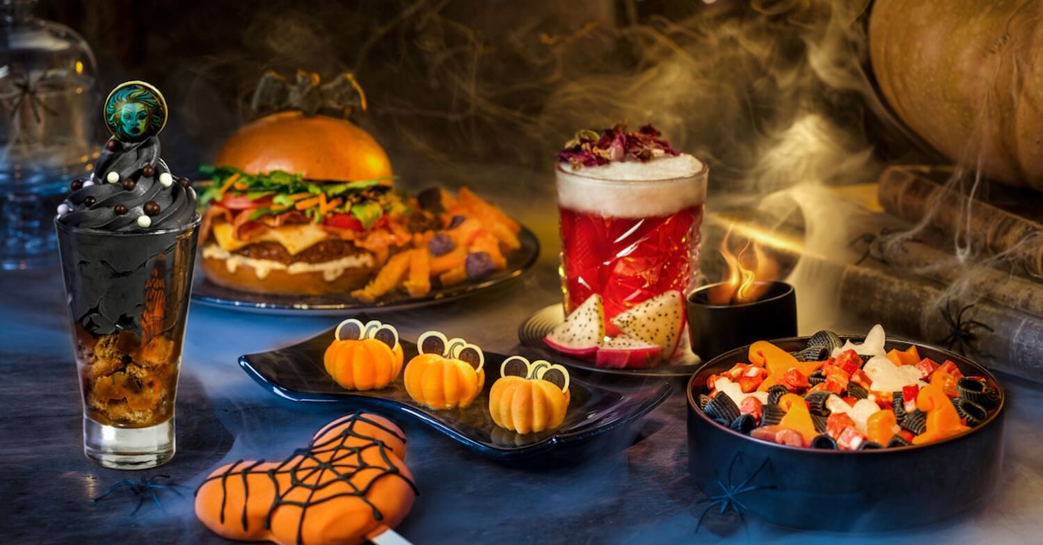 Snacks und Getränke im Halloweenlook sind zur Präsentation angerichtet. Orangene Kürbisküchlein in der mitte des Bildes, davor ein großer Mickey-Mouse Lolli mit Spinnennetz, ein rot dampfender Cocktail, ein Burger und andere orangefarbene Speisen