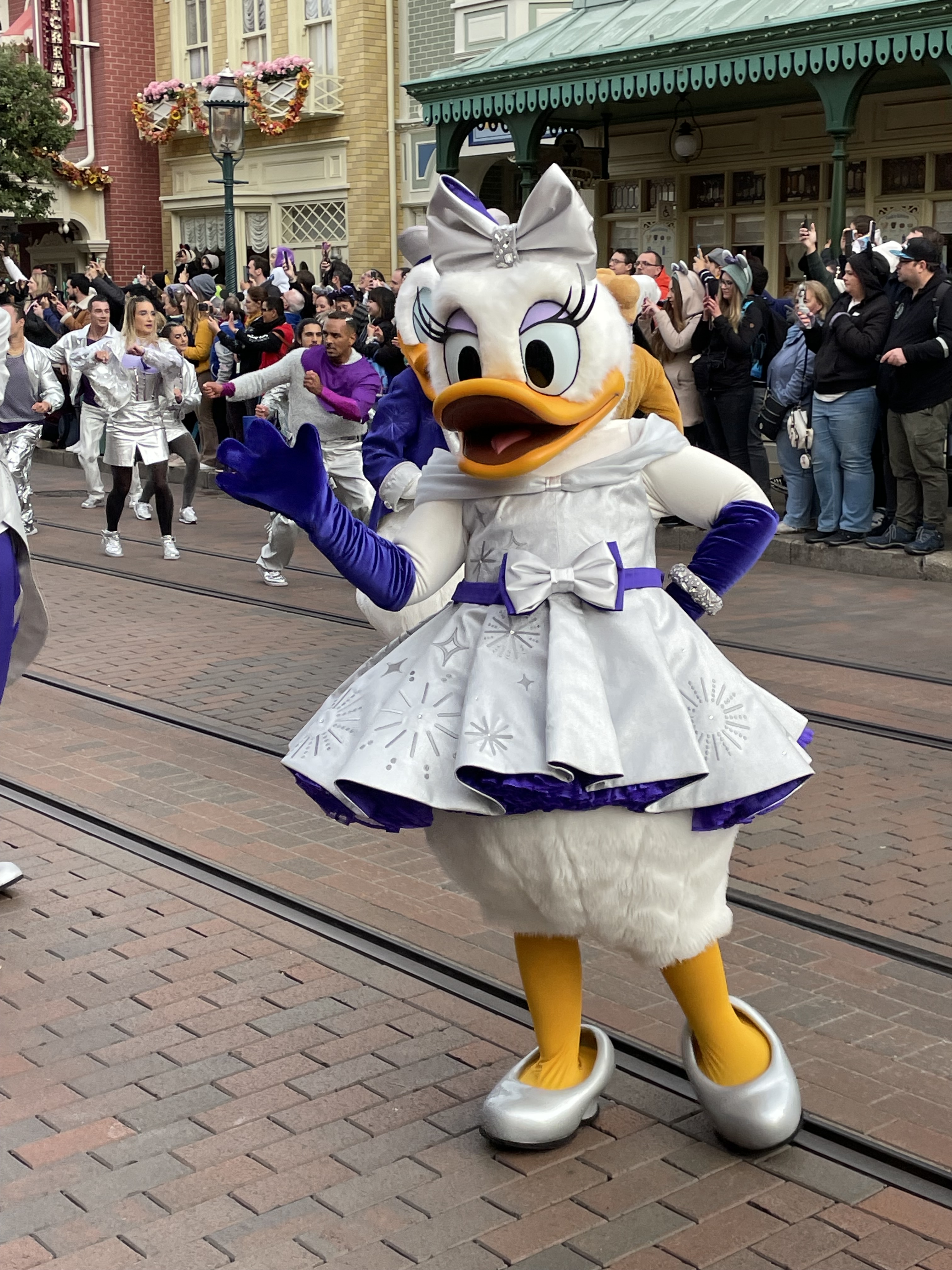 Daisy Duck trägt ein weißes Kleid mit silbernen Applikationen und lilafarbene Schmuckelementen. Sie winkt den Zuschauern zu