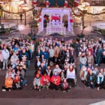 Eine Gruppe Menschen bildet die Form der Zahl 100 nach. Sie steht dazu auf dem beleuchteten Town Square in Disneyland Paris