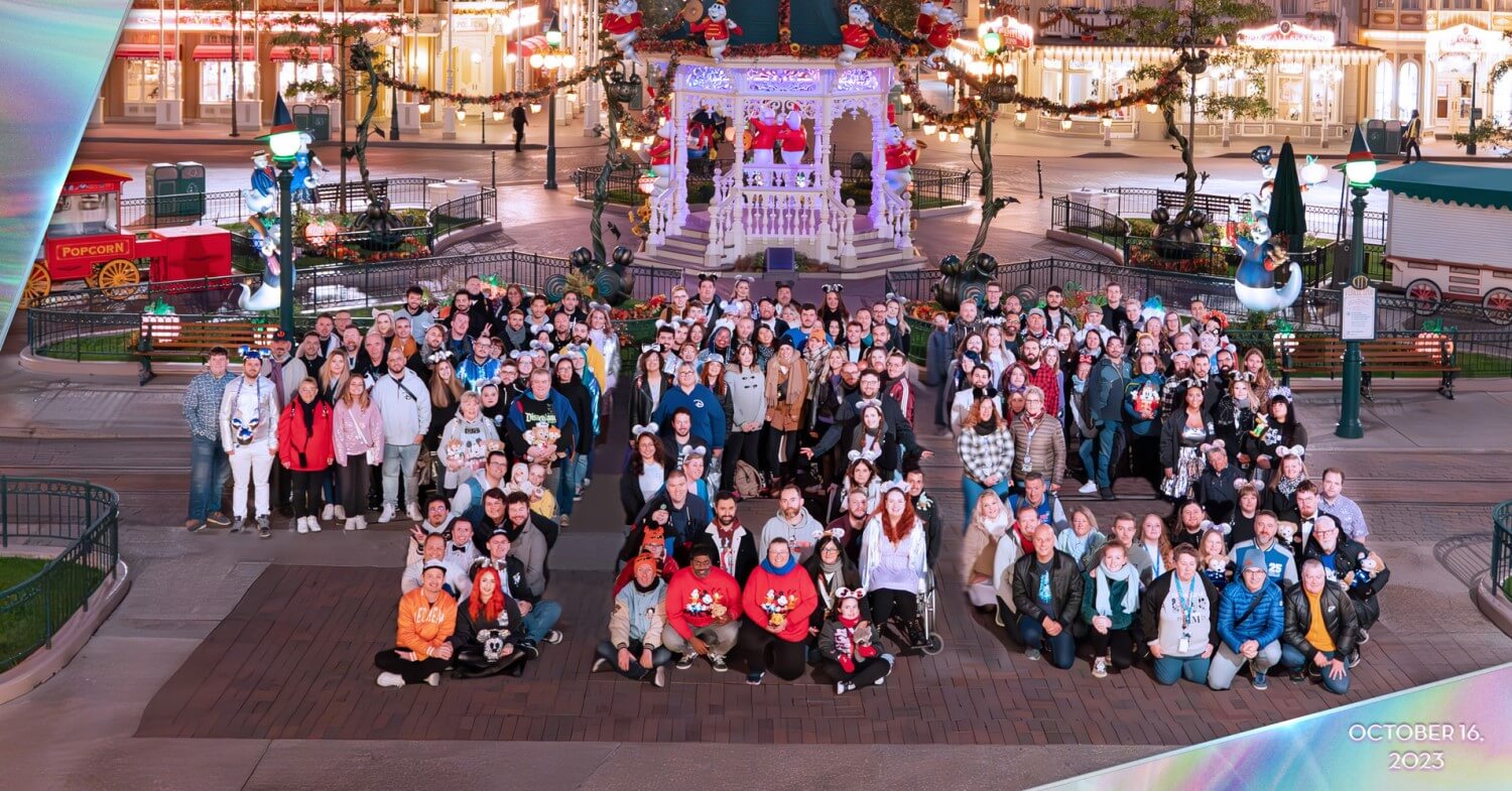 Eine Gruppe Menschen bildet die Form der Zahl 100 nach. Sie steht dazu auf dem beleuchteten Town Square in Disneyland Paris