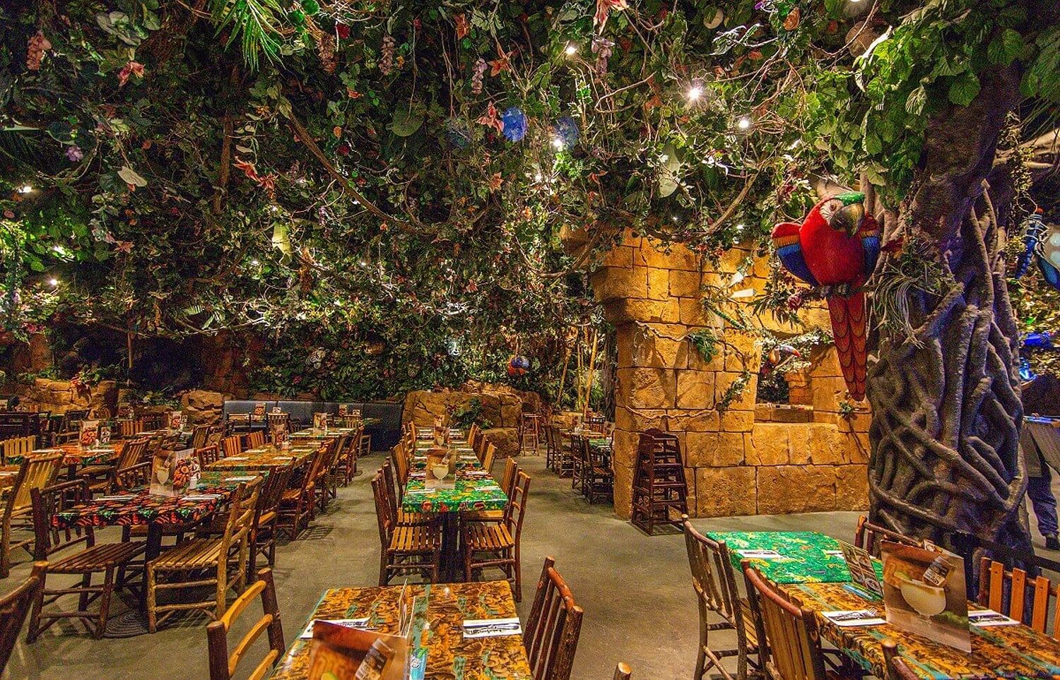 Dschungelatmosphäre im Gastraum des Rainforest Cafe. Tische und Stühle sind in Reihen angeordnet, an der Decke erwecken Kunstpflanzen den Eindruck eines Dschungels.