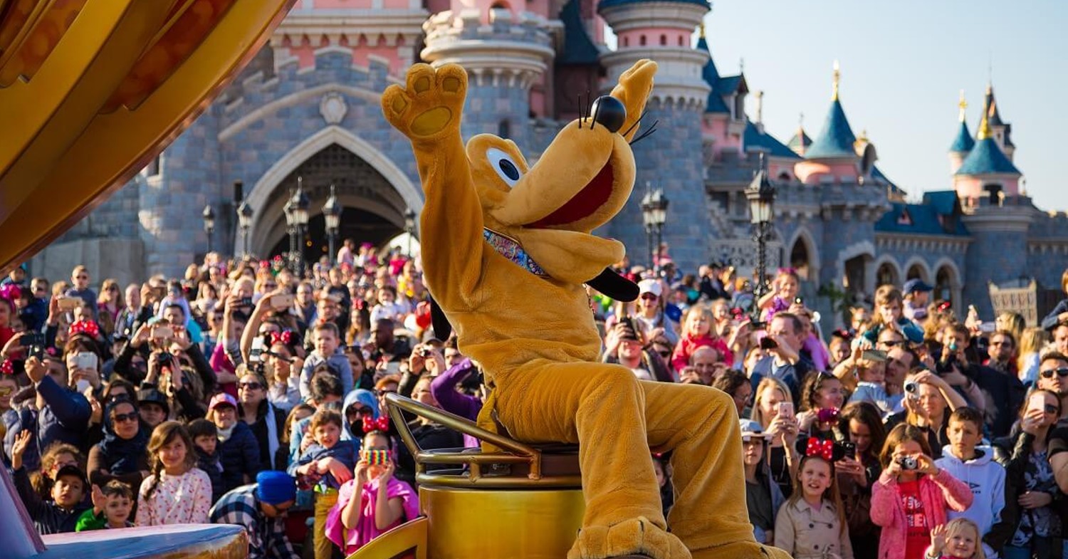 Pluto sitzt auf einem Paradenwagen, der gerade am Sleeping Bauty Castle vorbeifährt. Menschenmassen schauen ihm zu.