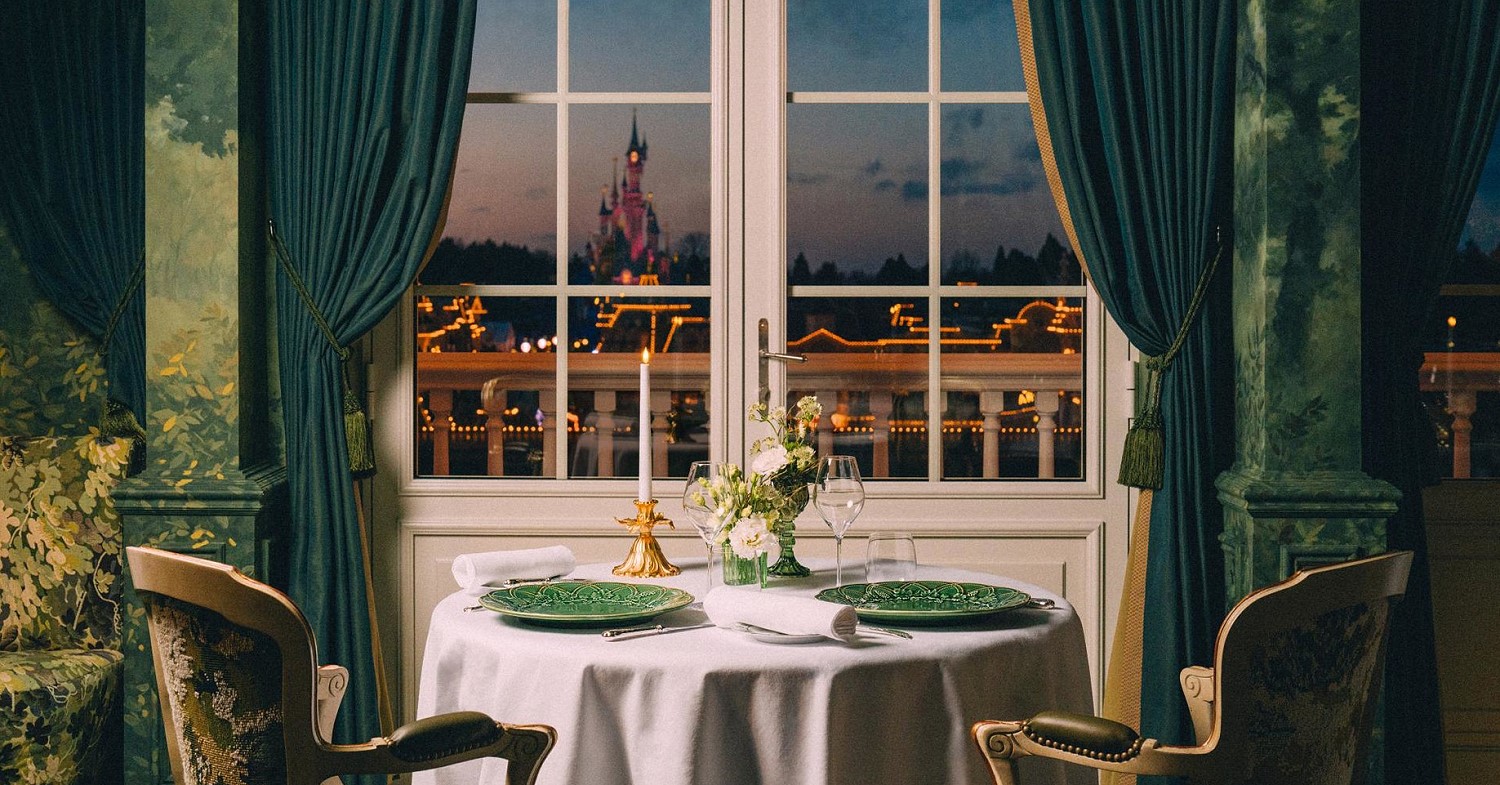 Gedeckter Tisch mit Stühlen vor einem Fenster, das Ausblick auf den Disneyland Park mit dem Sleeping beauty Castle bietet.